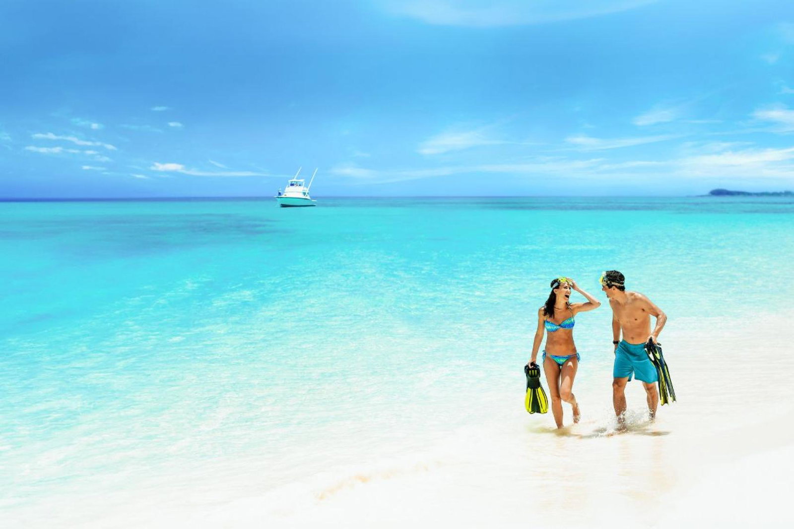 A young couple in swimwear walk along the white sand beach with their feet in the shallow of the blue sea