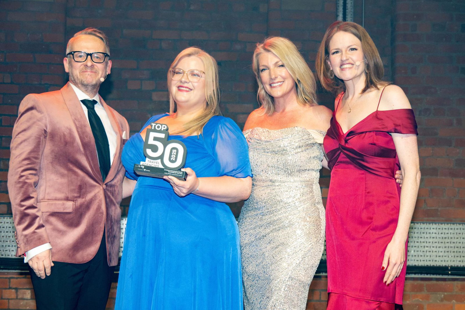 Four presenters stand on a stage in formal evening wear, smiling at the camera as the woman in a blue dress in the centre holds a “Top 50 Travel Agencies” award, with brick walls and stage lighting in the background.