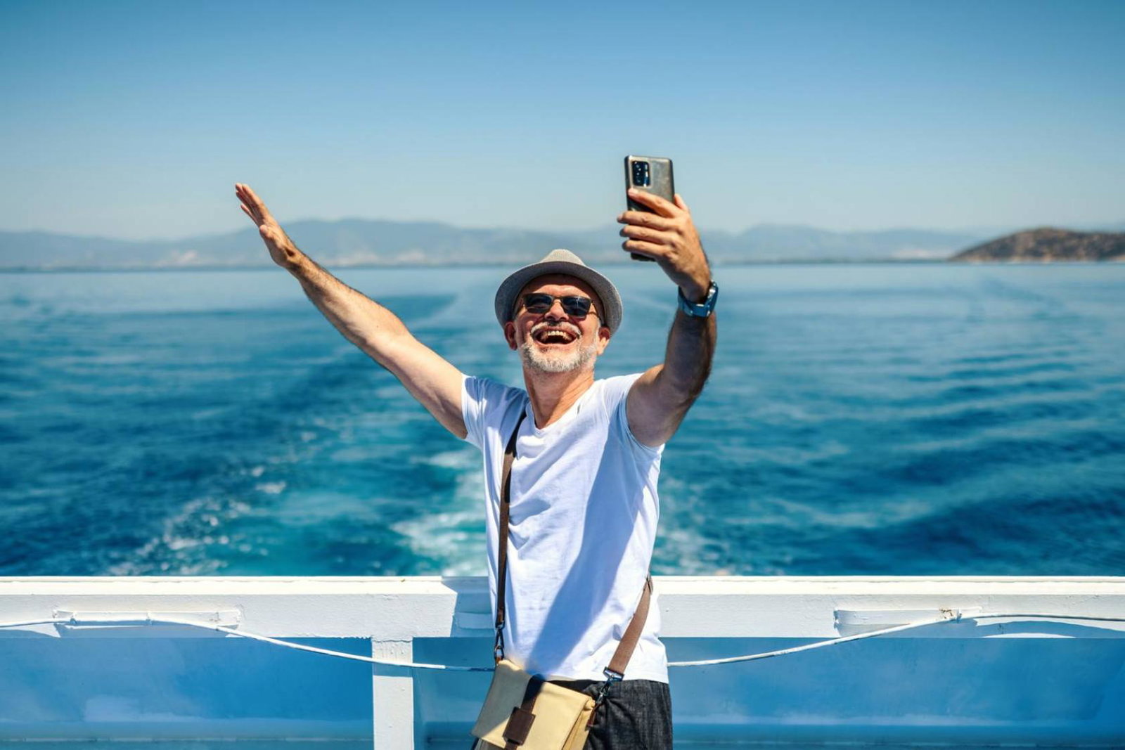 A mature man wearing a sunhat stands on the deck of a cruise ship, smiling and taking a selfie