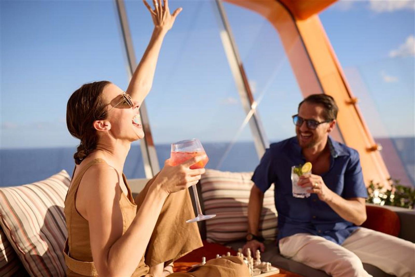 Two people sitting outside on a boat smiling with drinks, with a woman with her hand in the air