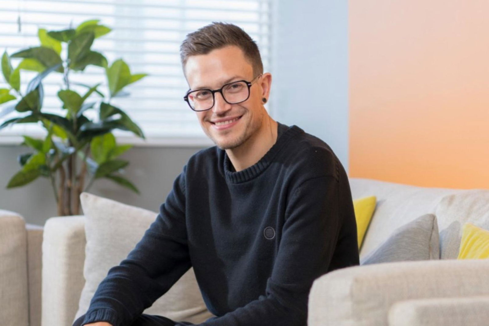 White man with glasses and short brown hair smiling, sitting on cream couch, plant in background