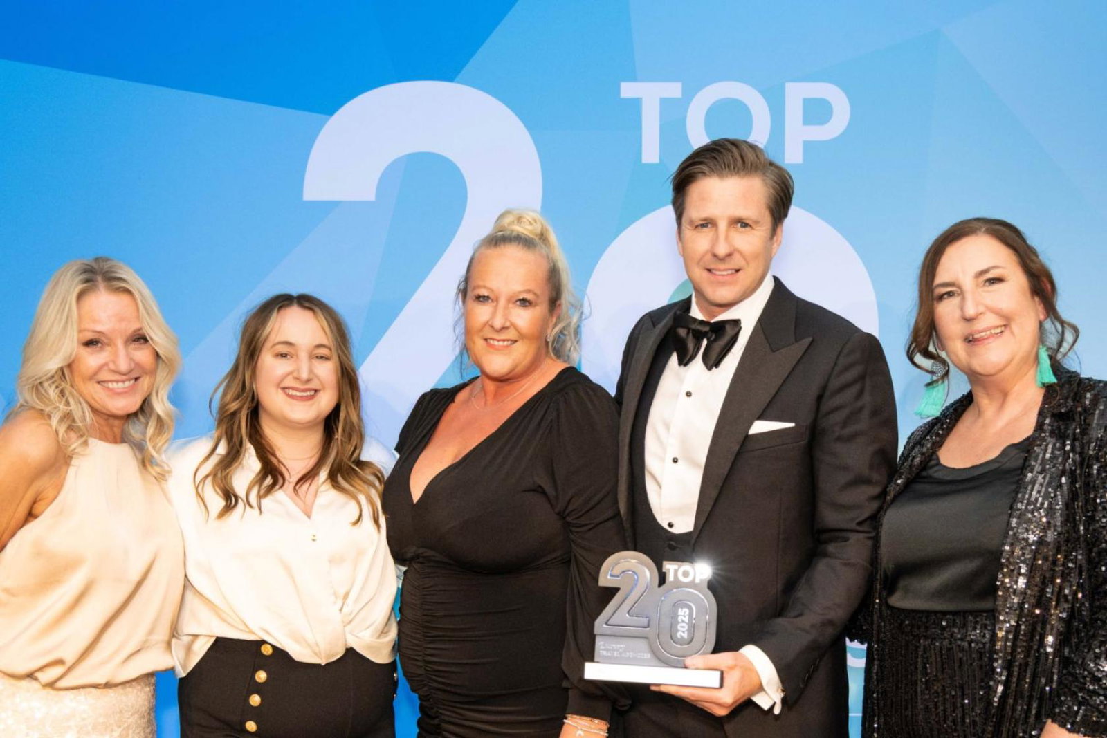 Four women smiling, one man smiling, man is holding trophy statue with 'Top 20' on it. Black tie event. 