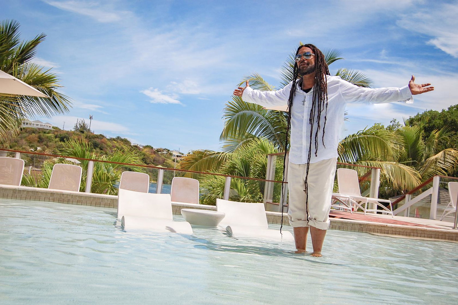 Casusion reggae artist in white clothing and sunglasses stands in a resort pool with palm trees, loungers and blue sky at The Verandah Antigua.