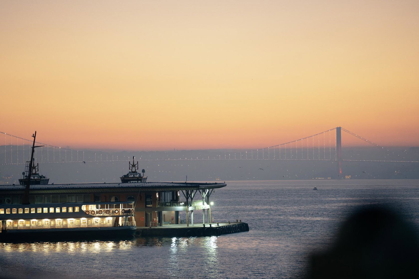 Istanbul’s waterfront and illuminated Bosphorus Bridge at sunset, with a ferry docked in the foreground.