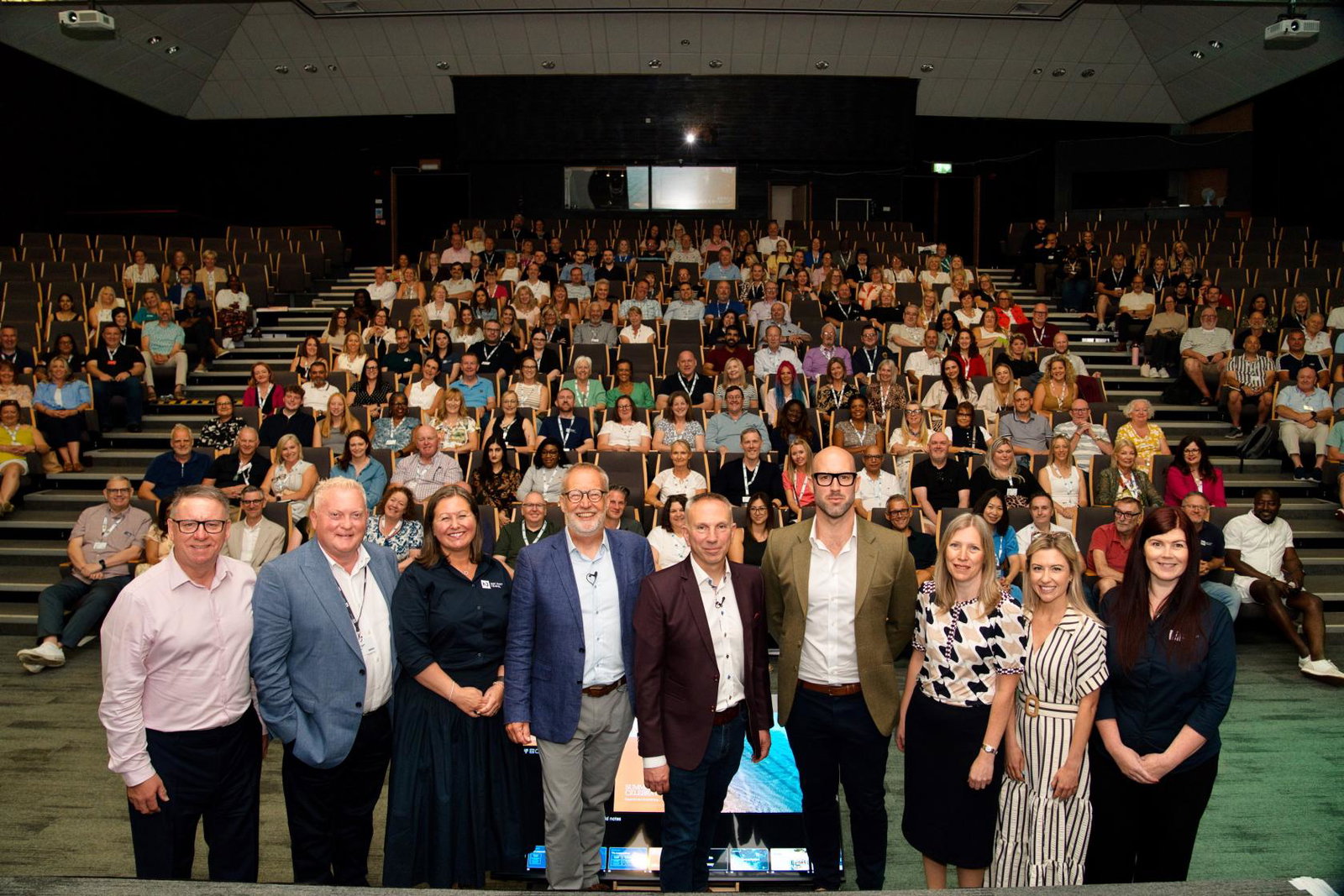 Not Just Travel corporate team: Dave Pope, Duncan Croucher, Emma Holliday, Derek Jones, Steve Witt, John Jones, Helen Foottit, Josie Allen, Emma Lawson in front of an auditorium of travel consultants at Summer Celebration