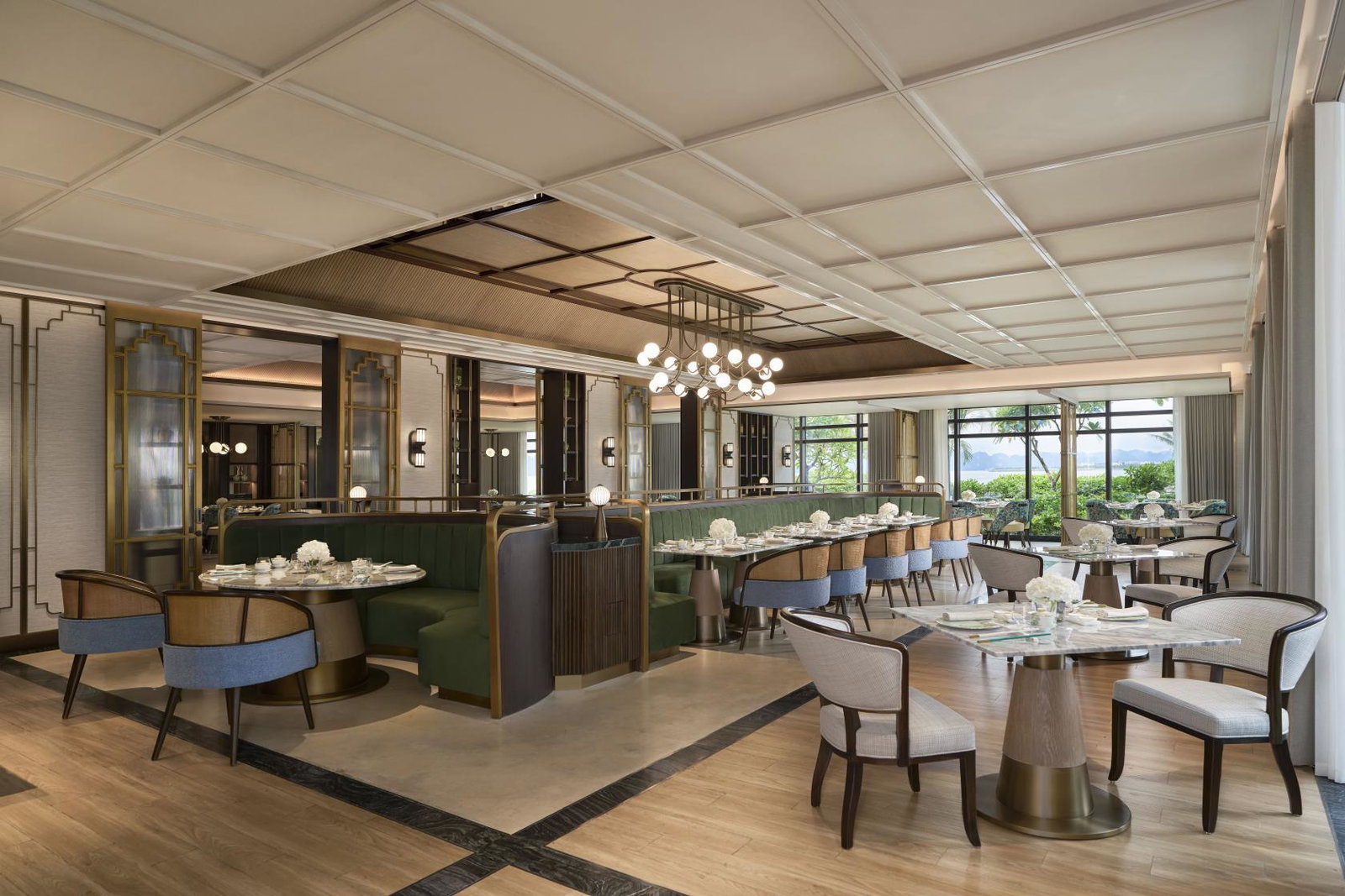 Elegant dining room at Yulong Mansion with green velvet booths, marble tables, and brass accents, with windows overlooking Ha Long Bay.