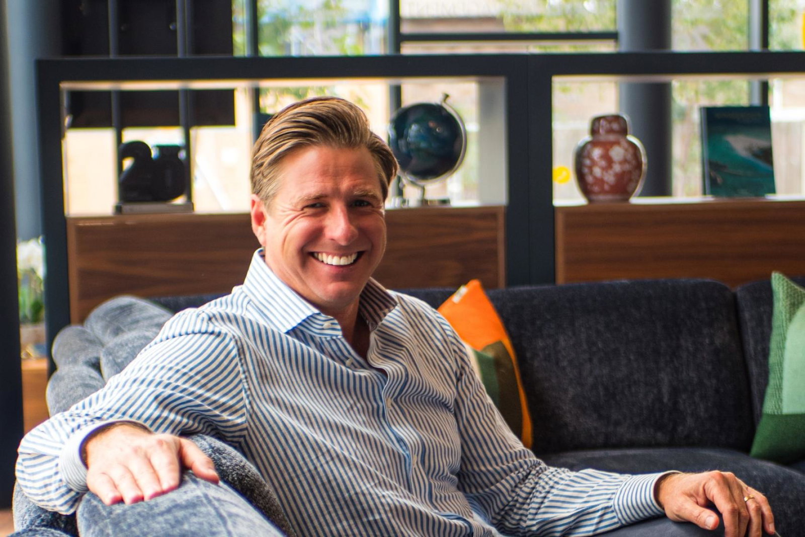 White man smiling, wearing blue shirt and sitting on sofa 
