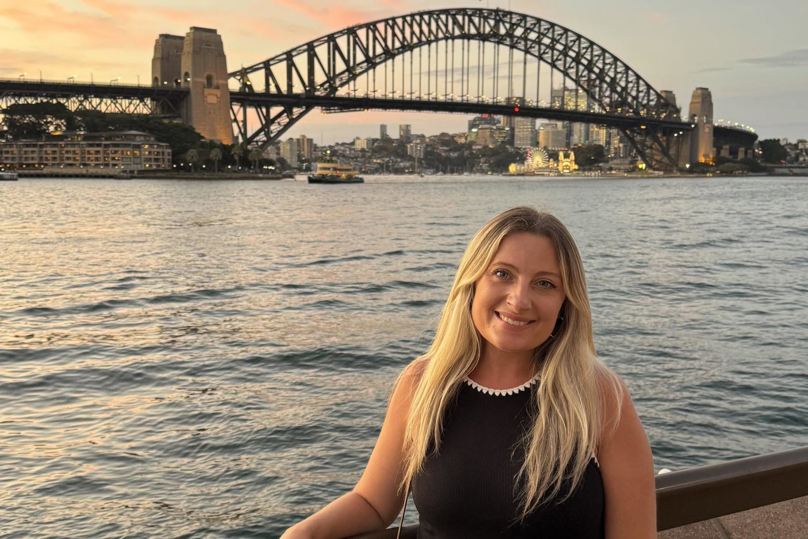 Chloe Seaforth, ANZCRO UK's South UK Sales Manager, smiling in front of the Sydney Harbour Bridge at sunset, with the harbour, ferries and Luna Park lit up behind her.