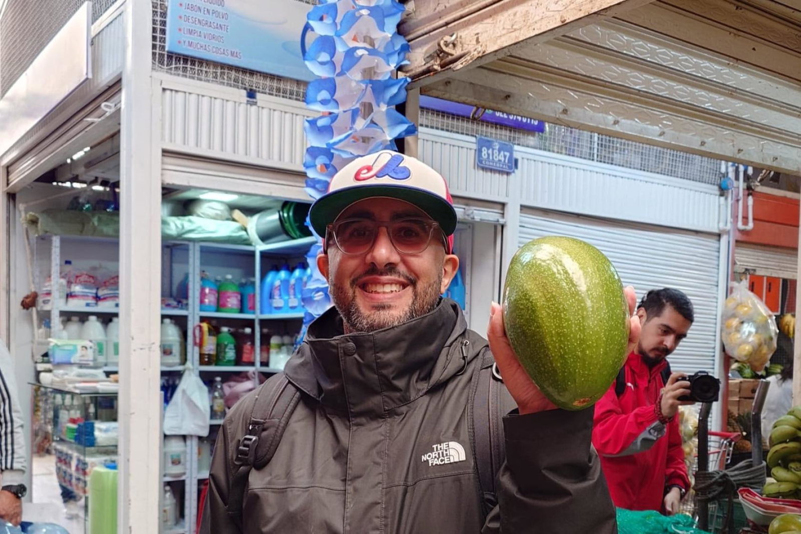 A smiling brown-skinned man with glasses and a baseball cap, holding up a large green fruit at a market