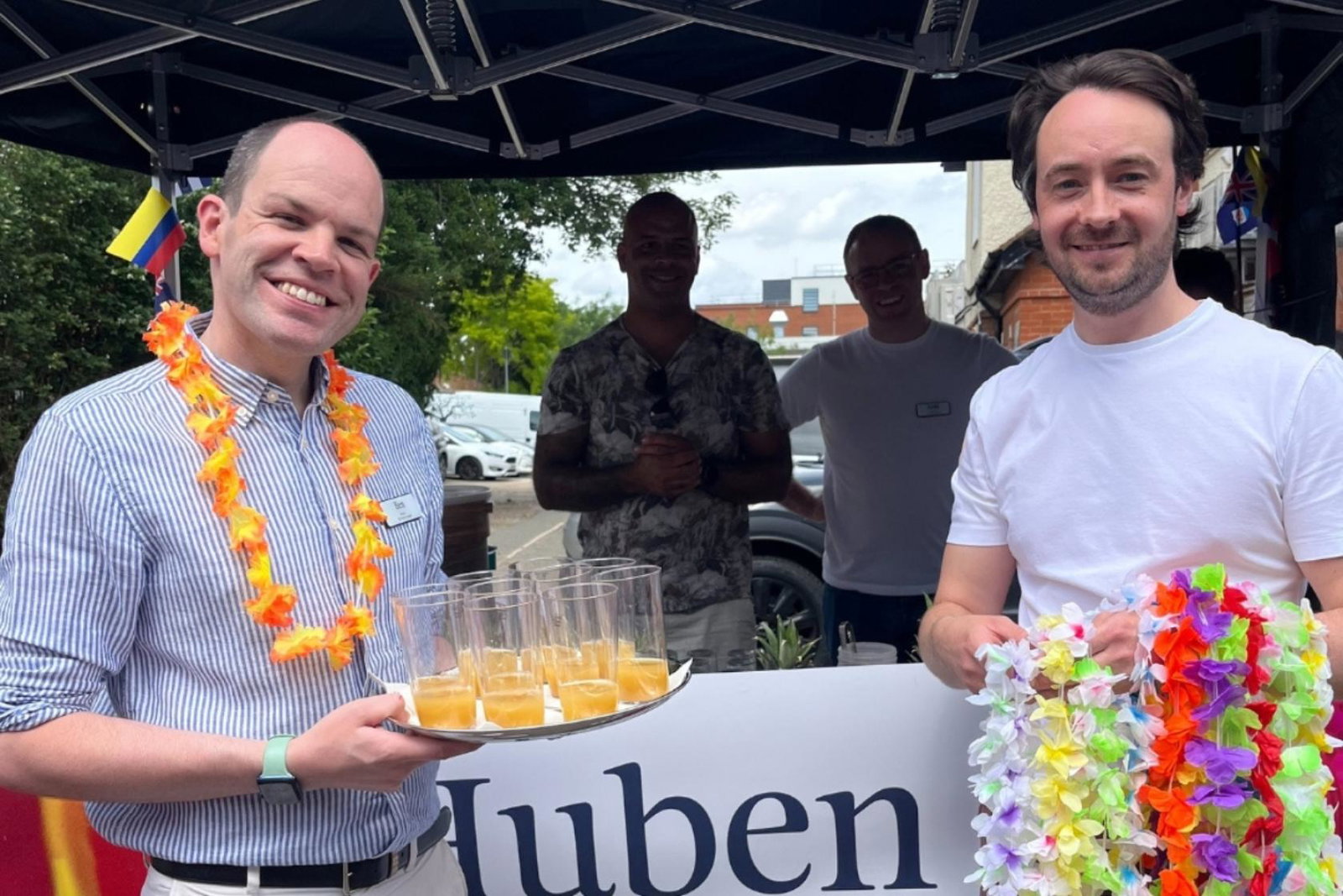 Two white men smiling in front of a stall and wearing necklaces made of flowers 