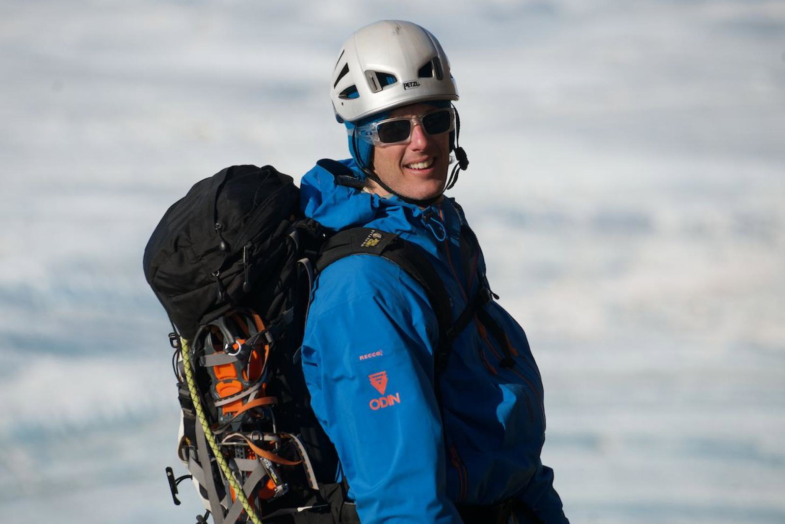 White man smiling in Antarctica, wearing helmet and sunglasses and blue jacket  