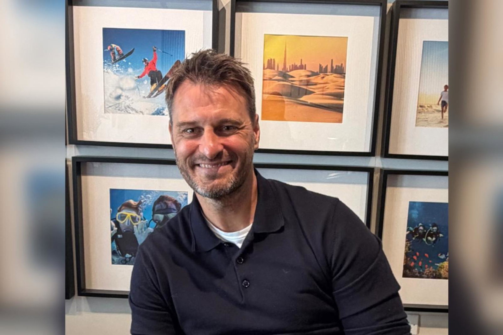 White man smiling with framed travel-themed pictures behind him  