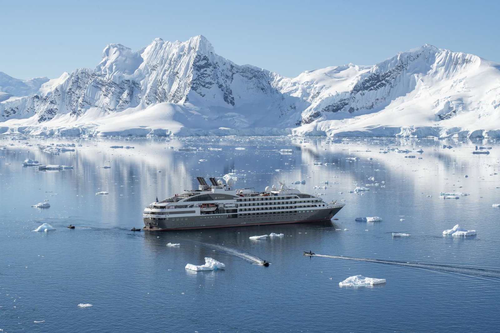 PONANT's ship L'Austral anchored among floating ice in calm blue waters, with Zodiac boats heading out and snow-covered mountains reflected in the sea behind.