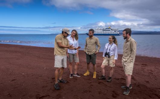 Guide and tour group on red sand beach with ship in the distance