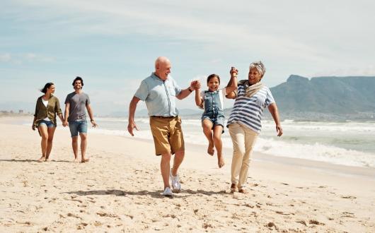 Grandparents play with their grand-daughter on the beach while parents walk behind