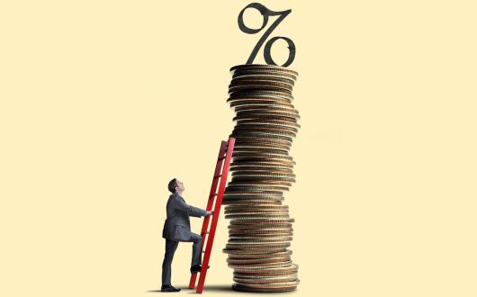 A man looks up as he leans a red ladder against a tall stack of coins that is topped with an interest rate symbol.