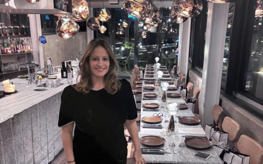 Sara Greene, brown-haired smiling woman standing in front of long dining table 