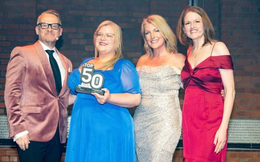 Four presenters stand on a stage in formal evening wear, smiling at the camera as the woman in a blue dress in the centre holds a “Top 50 Travel Agencies” award, with brick walls and stage lighting in the background.
