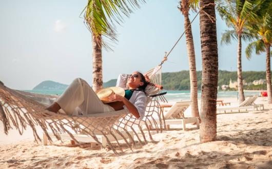 A woman relaxes on a hammock in a tropical beach location with palm trees