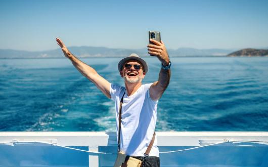 A mature man wearing a sunhat stands on the deck of a cruise ship, smiling and taking a selfie