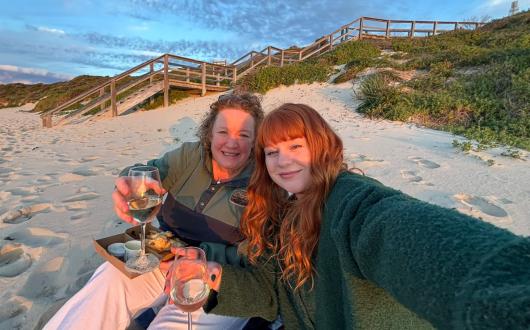 Mum and adult daughter, both with red hair, sitting on a sandy beach and holding glass of white wine 