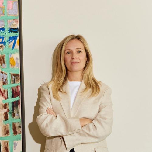 Blonde woman standing in front of white wall, to the left is a painting 