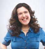 A white woman with dark hair, the comedian Rosie Jones, is standing against a white backdrop. She is smiling and wearing a blue shirt 