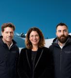 A professional image of a white smiling woman standing with two white men on each side of her, background is snowy ski slopes 