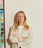 Blonde woman standing in front of white wall, to the left is a painting 