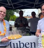 Two white men smiling in front of a stall and wearing necklaces made of flowers 