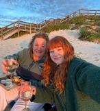 Mum and adult daughter, both with red hair, sitting on a sandy beach and holding glass of white wine 