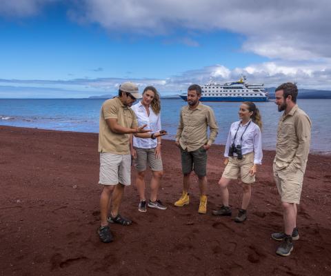 Guide and tour group on red sand beach with ship in the distance