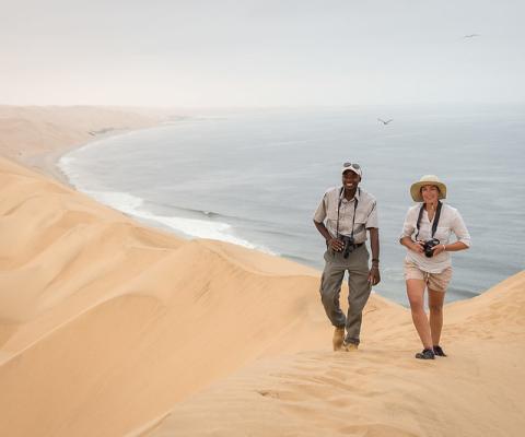 People walking over giant sand dunes with ocean behind