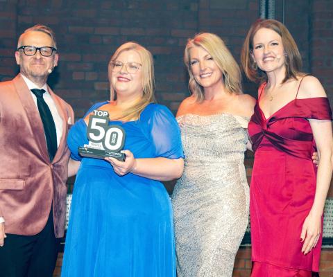 Four presenters stand on a stage in formal evening wear, smiling at the camera as the woman in a blue dress in the centre holds a “Top 50 Travel Agencies” award, with brick walls and stage lighting in the background.
