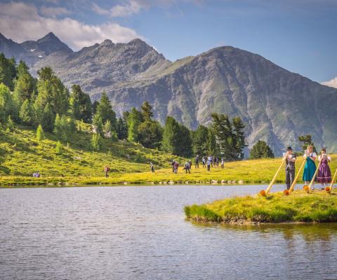 Landscape with lake, mountains and trees with three people playing traditional alpine horns