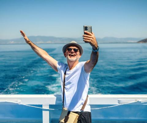 A mature man wearing a sunhat stands on the deck of a cruise ship, smiling and taking a selfie