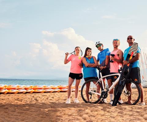 Group of people on beach holding different sporting equipment, from bikes to tennis racquets