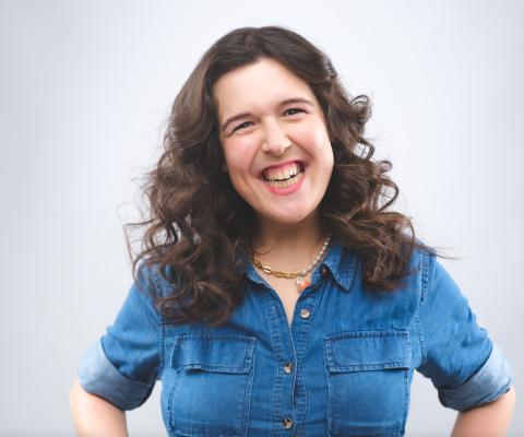 A white woman with dark hair, the comedian Rosie Jones, is standing against a white backdrop. She is smiling and wearing a blue shirt 