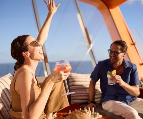 Two people sitting outside on a boat smiling with drinks, with a woman with her hand in the air