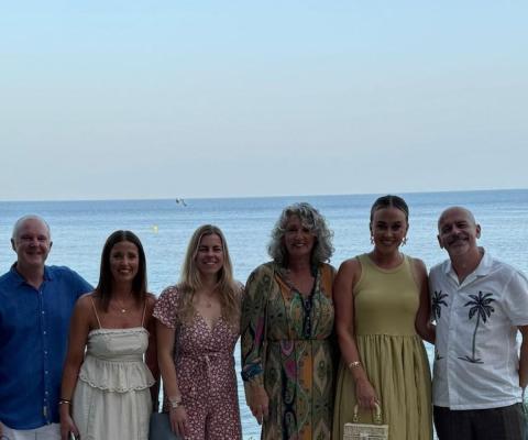 Six people, two men and four women, smiling in front of a beach 