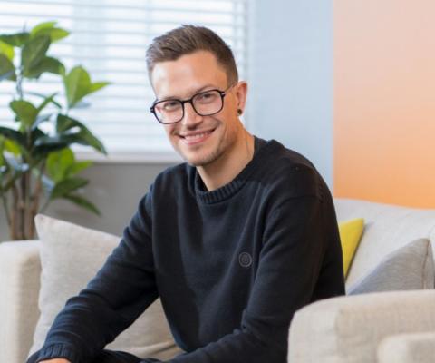 White man with glasses and short brown hair smiling, sitting on cream couch, plant in background