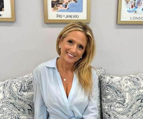 Woman with long blonde hair smiling, wearing light blue shirt, framed photographs in the background