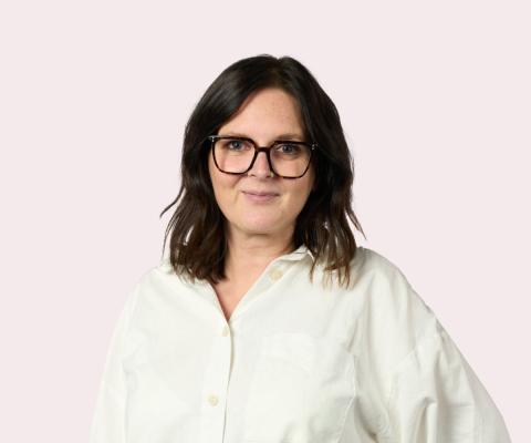 Headshot of brown haired woman with glasses and white shirt, light pink background,