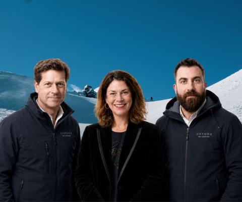 A professional image of a white smiling woman standing with two white men on each side of her, background is snowy ski slopes 