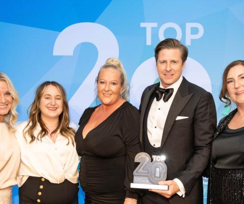 Four women smiling, one man smiling, man is holding trophy statue with 'Top 20' on it. Black tie event. 