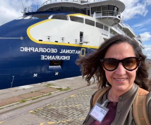 Woman with brown long hair and sunglasses smiling in front of blue and white ship 