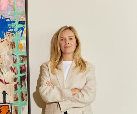 Blonde woman standing in front of white wall, to the left is a painting 