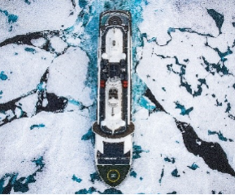 Aerial view of an expedition cruise ship navigating through fractured sea ice in polar waters, its dark hull cutting a path through a mosaic of white ice floes and dark blue-green water below.