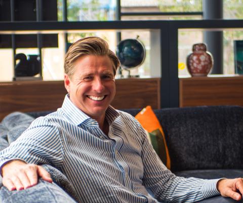 White man smiling, wearing blue shirt and sitting on sofa 