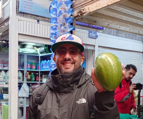 A smiling brown-skinned man with glasses and a baseball cap, holding up a large green fruit at a market